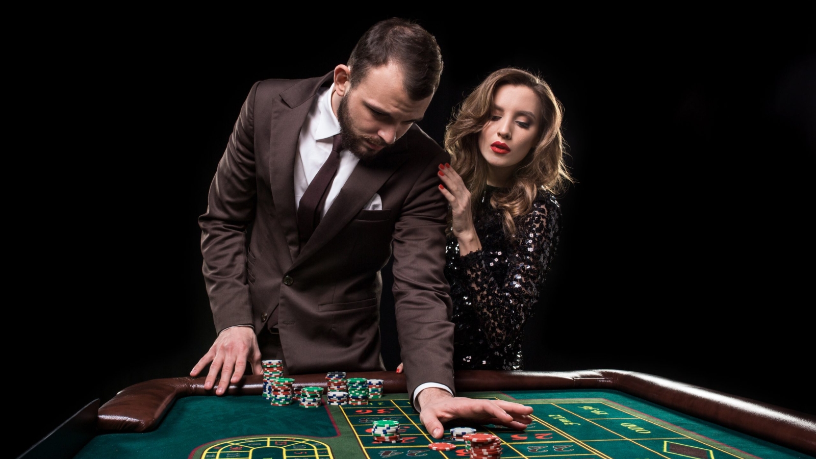 A man in a brown suit with a beautiful woman in a black dress is playing at a roulette table at a casino. Emotion of gambling players