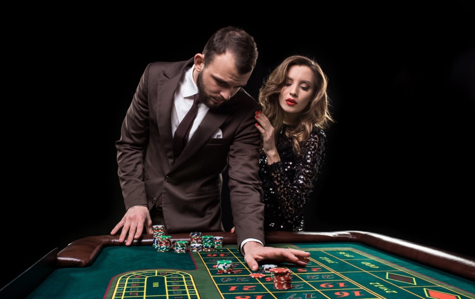 A man in a brown suit with a beautiful woman in a black dress is playing at a roulette table at a casino. Emotion of gambling players